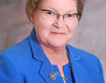 Older woman with short light brown hair and glasses in a blue suit.