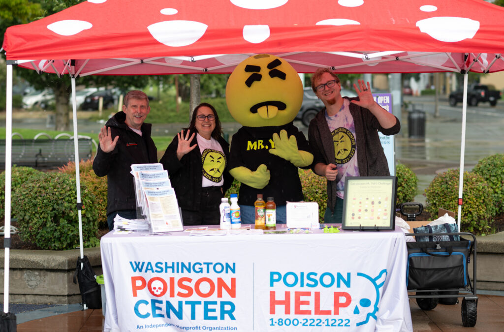 Mr. Yuk Mascot - a green character with a round head, disgusted look and sticking-out tongue), two men and a woman standing under a red and white polka-dotted tent behind a table with a poison help tablecloth waving