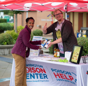A man in his 30s at the Washington Poison Center tent giving a thumbs up while talking to a black woman, also looking at the camera
